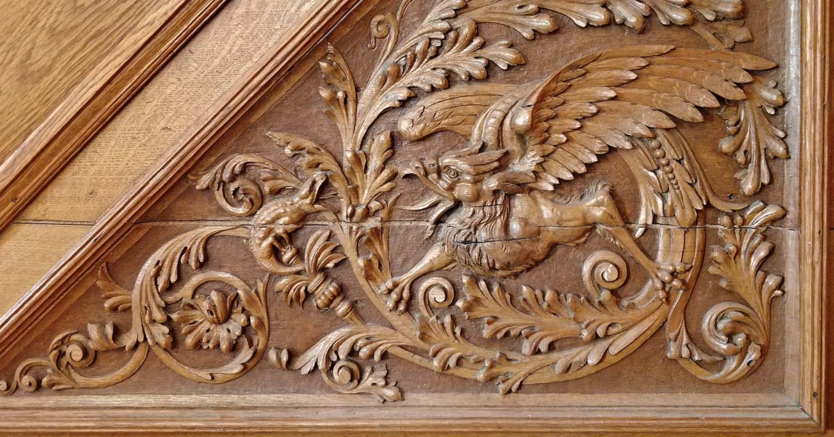 wood panel carving depicts a gryphon (mythical eagle/lion) attacking a lizard, surrounded by decorative vines
