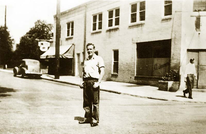 Cafe building's Iona side; young man stands in street looking at camera; an old car is parked on curb.