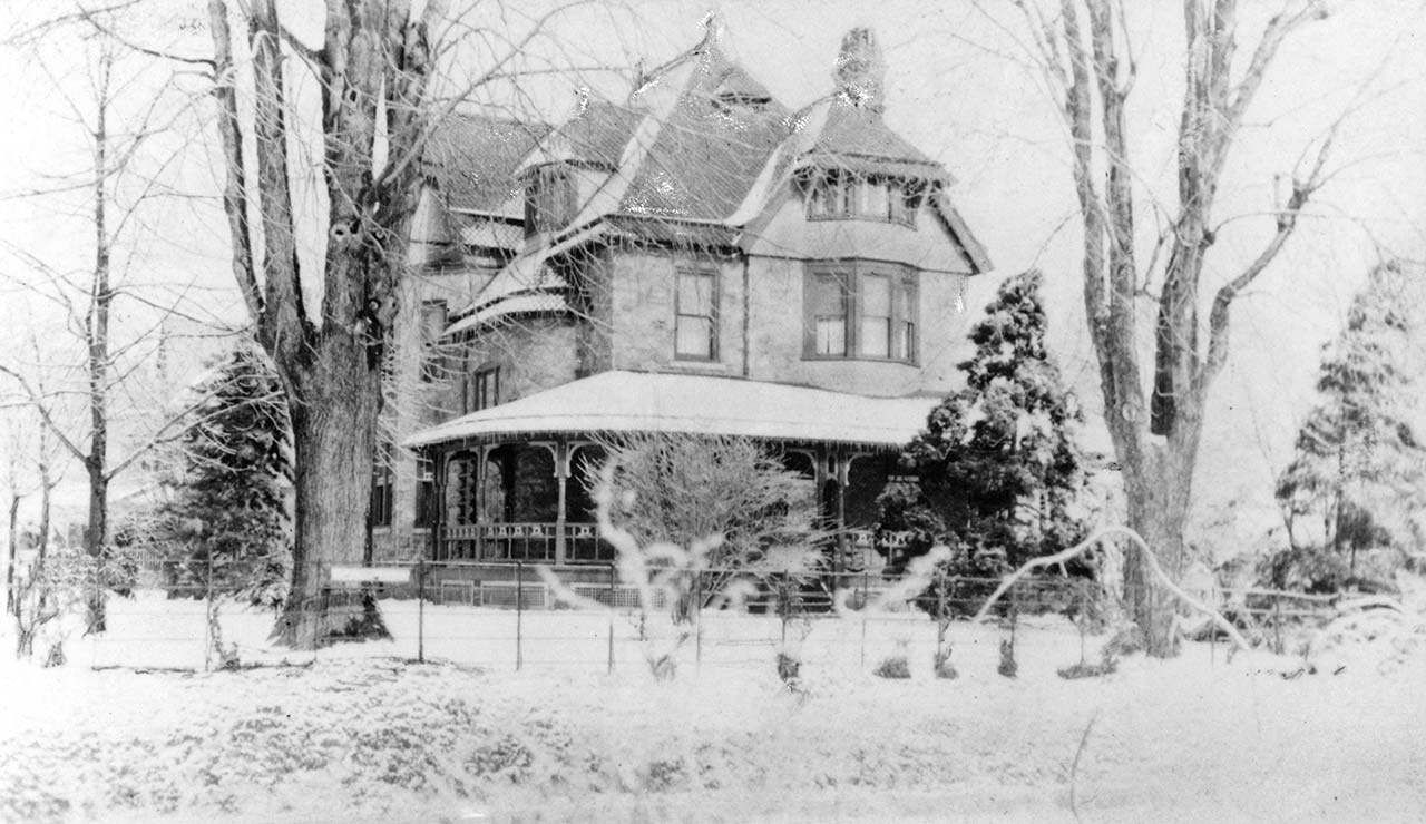 large stone house with wrap-around porch in a snowy yard with 2 big bare trees