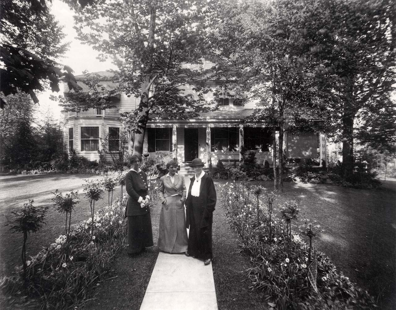 three women in ankle-length dresses stand on walway leading to a large house. There are formal  plantings on either side of the walkway.