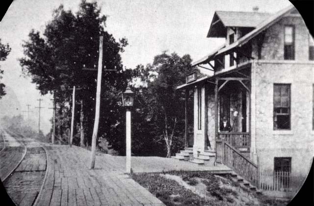 looking down a two-track rail line; beside a platform of wooden planks with a lampost and a three-story stone station house.