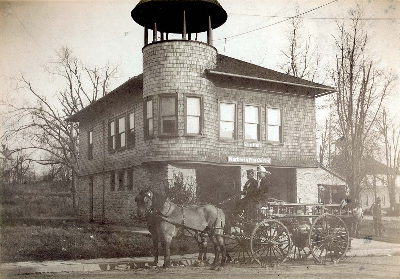 A horse-drawn firewagon driven by two firemen stands in front of the firehouse.