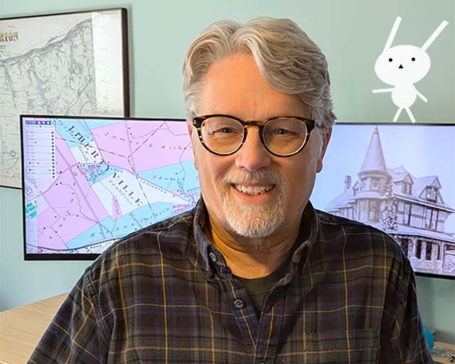 standing in front of screens showing 1871 map and Victorian turreted house; Narbunny is perched on edge of monitor