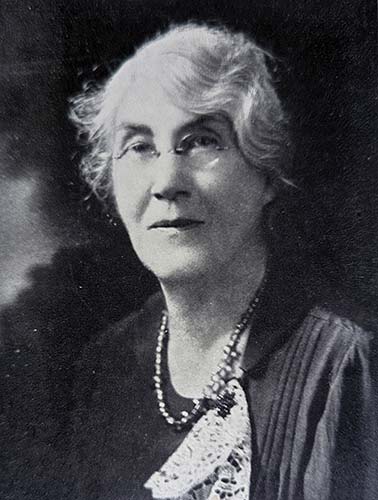 head and shoulders portrait of a woman with short gray hair, round wire glasses, pearl necklace