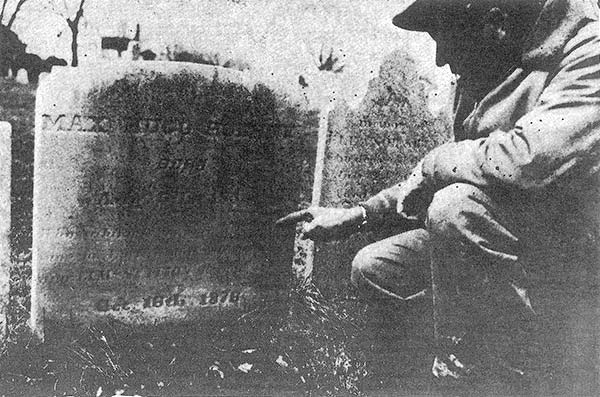 man squats and points at an old mostly illegible grave stone that read 'Max Hugo Hoehne'