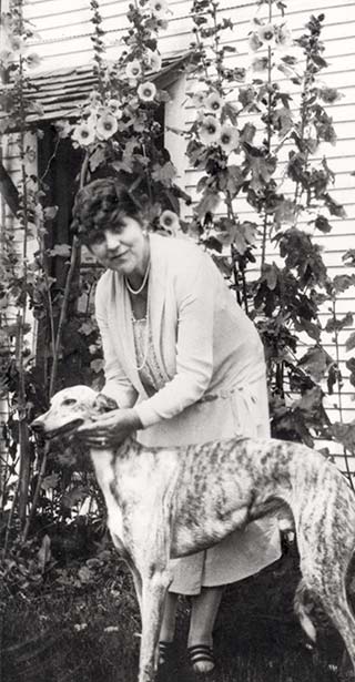 A middle-aged woman standing in front of stand of hollyhocks, pets a racing dog