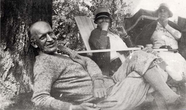 man wearing tie, buttoned sweater and knickers lies on ground against a tree, with two women in Adirondack chairs