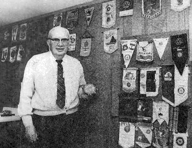 older balding man holding a baseball standing in front of wall covered in banners