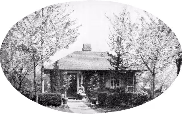 one-story cottage surrounded by small flowering trees; a man with a child on his lap sit by its door
