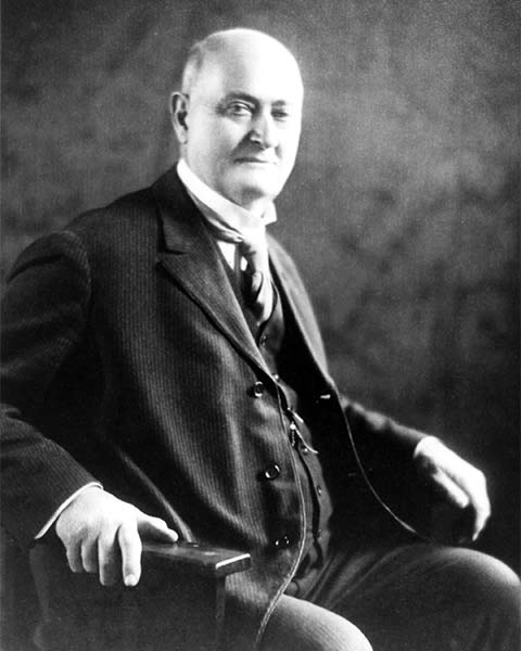 older bald man seated, in 3-piece suit, watch chain