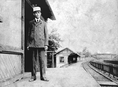 smartly uniformed man standing in doorway on train platform; tracks and Narberth Ave. bridge in background.