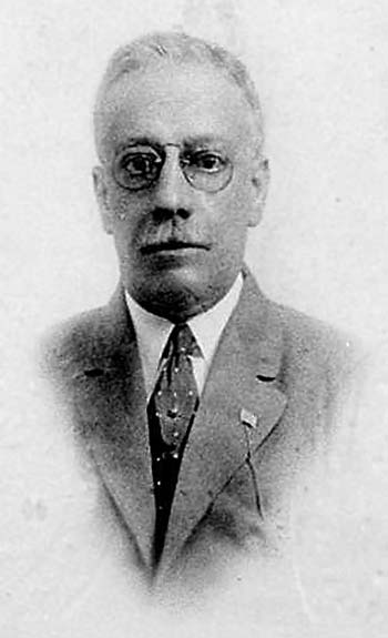 grainy head and shoulders of older man in suit with mustache, wireframe glasses, lapel pin