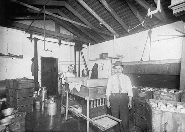 2 men stand among equipment, milk cans and milk bottles in wooden boxes