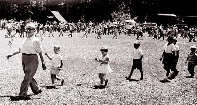 older man leads a line of children in the park, all holding flags