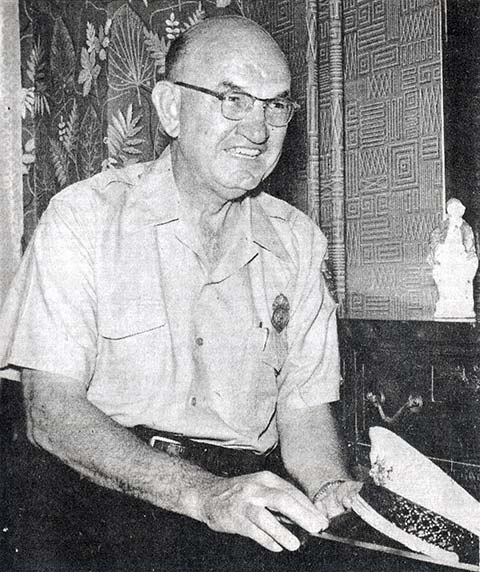 smiling policeman seated in short sleeved uniform, holding police cap