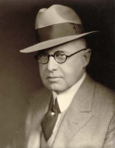 formal head shot of a middle aged man in 3-piece suit and tie, fedora hat, round-frame glasses