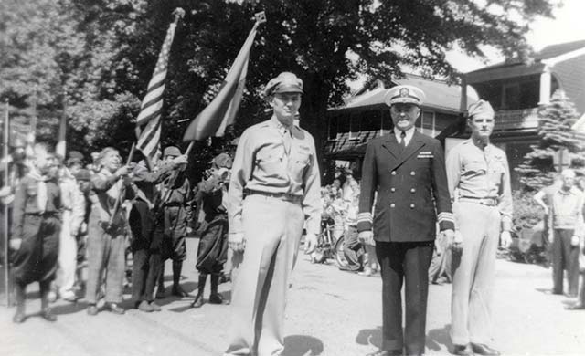 Commander O'Sullivan in a dark dress uniform stands at attention, flanked by his two sons in uniform; behind them lines up an enthusiastic boy scout group bearing 2 flags