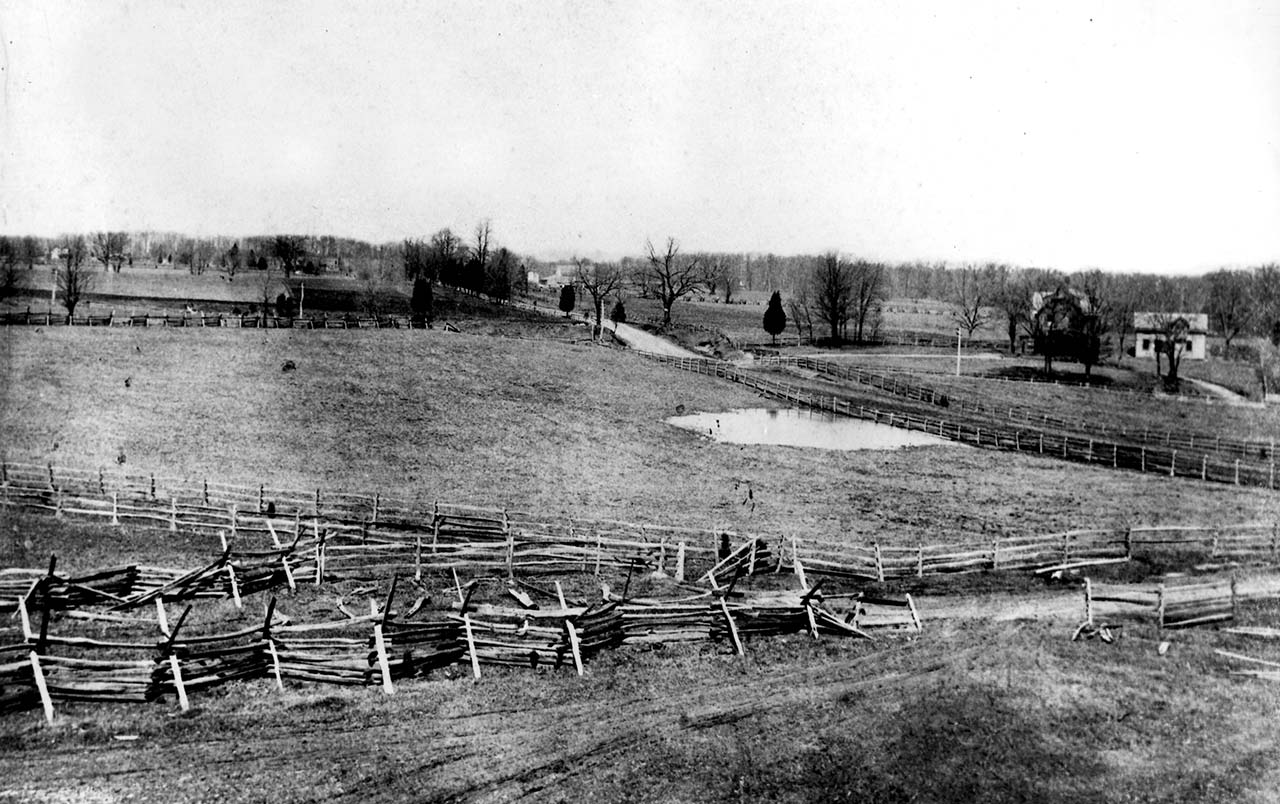 A landscape of open rolling fields, crisscrossed by rail fences, dirt roads and wagon paths, a few scattered houses and bare winter trees in the background.