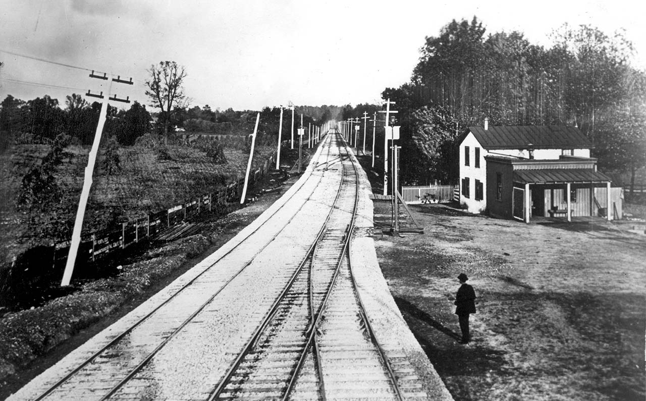 Two train tracks on a gravel bed with telegraph poles on both sides recede into the background. A field with sheaves of corn is to the left and a small two-story house on the right. A man stands beside a siding looking down the tracks.