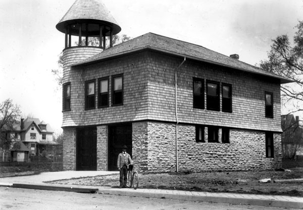 A Black man holding a bicycle stands on a paved sidewalk by a dirt street in front of a building with a stone first story and two full-height doors. A circular open watchtower capped by a pointed turret rises above the corner of its second story.