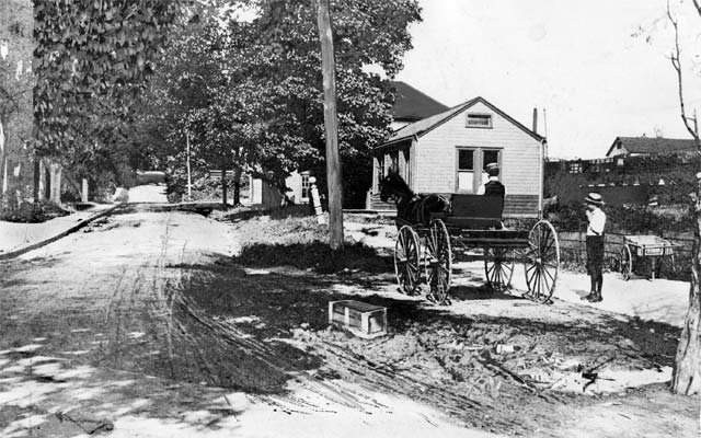 boy in knickers next to a horsecart with a seated driver, standing on a sidewalk beside a muddy lane overshaded by large leafy trees