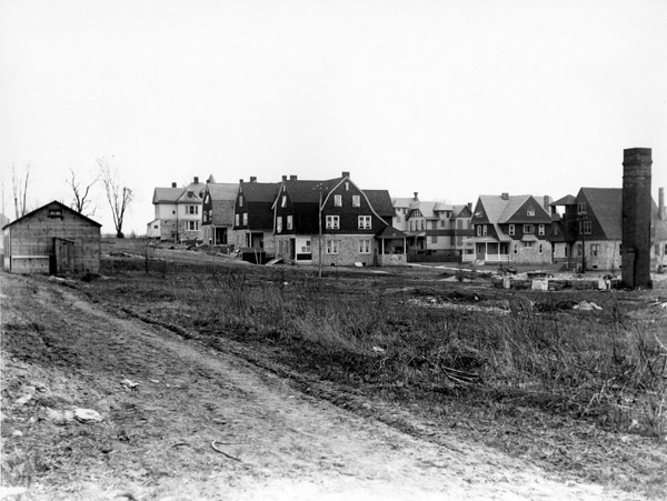 An empty lot save for an isolated chimney and some stone foundations. In front, a small wood building beside a dirt road. In the background, a block of closely spaced three-story houses.