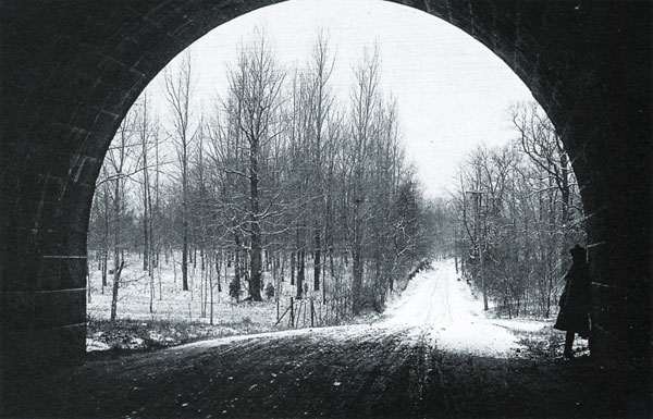 Looking out of a circular arched stone tunnel on a snowy dirt road that runs through woods. A silhouetted figure leans against the tunnel wall.