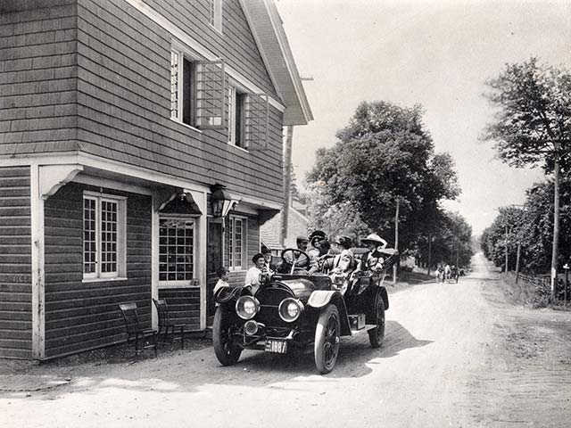 Well-appointed antique car with five well-dressed occupants stops in front of 3-story house on unpaved road; they are attended by a man, woman and boy. A horsecart approaches on unpaved, wooded Montgomery Ave.