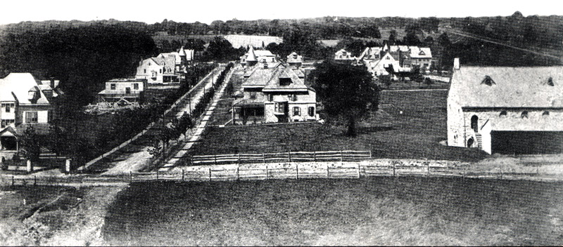A roof-level view over south Narberth showing a dirt crossroads in the foreground, a dozen or so late Victorian houses, one under construction, and a stone church with a wood shed.
